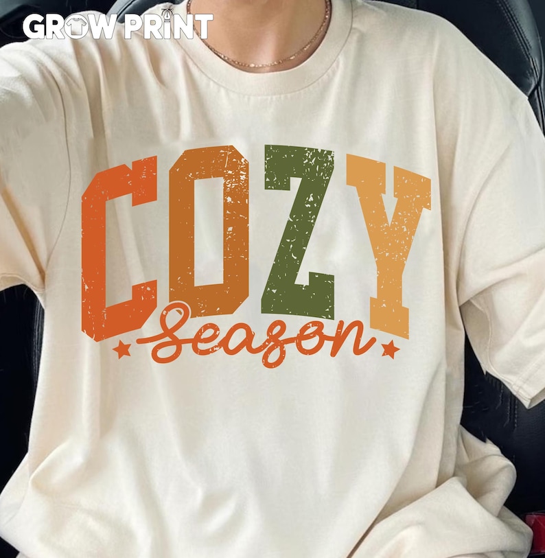 Peut inclure: T-shirt cr&egrave;me avec l'inscription "COZY Season" en lettres d&eacute;lav&eacute;es orange, vert et jaune. Le t-shirt a un col rond et des manches courtes. La marque "GROW PRINT" est en haut.