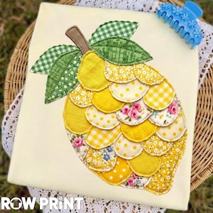 Puede incluir: Una aplicación decorativa de limón sobre tela color crema. El limón está hecho de piezas de tela amarilla con varios patrones, incluyendo lunares, estampados florales y cuadros vichy. Hojas verdes y un tallo marrón completan el diseño. Una pinza azul para el pelo descansa sobre la tela.