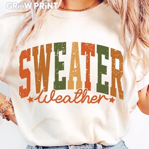 Peut inclure: T-shirt cr&egrave;me avec l'inscription "SWEATER Weather" dans une police r&eacute;tro vieillie. Le texte est dans des tons orange, vert et marron, avec des &eacute;toiles. Le t-shirt a un col rond.