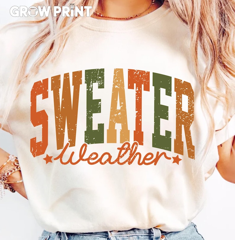 Peut inclure: T-shirt cr&egrave;me avec l'inscription "SWEATER Weather" dans une police r&eacute;tro vieillie. Le texte est dans des tons orange, vert et marron, avec des &eacute;toiles. Le t-shirt a un col rond.