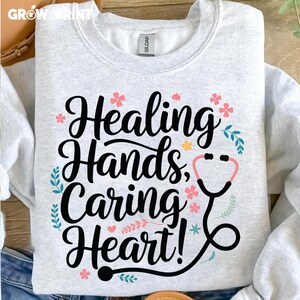 May include: A light gray sweatshirt with the text "Healing Hands, Caring Heart!" in black lettering. The text is surrounded by small pink and yellow flowers and green leaves. A black stethoscope with a pink circle for the listening end is also included in the design.