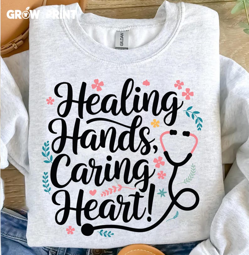 May include: A light gray sweatshirt with the text "Healing Hands, Caring Heart!" in black lettering. The text is surrounded by small pink and yellow flowers and green leaves. A black stethoscope with a pink circle for the listening end is also included in the design.