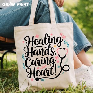 May include: A white canvas tote bag with black text that reads "Healing Hands, Caring Heart!" and a black stethoscope graphic. The bag has pink and blue floral accents.