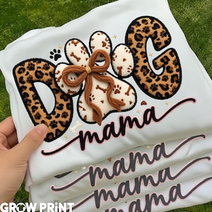 May include: White sweatshirt with the words "DOG mama" in a combination of leopard print and embroidered paw prints. The design includes a brown bow and bone accents. The sweatshirt is folded, with the text "mama" repeated multiple times.