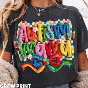May include: Dark gray t-shirt with the words "AUTISM MAMA" in colorful, textured letters. The design includes a rainbow bow and puzzle piece accents, with a checkered background. The shirt is a casual style.