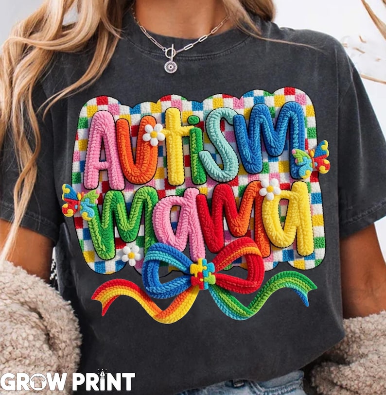 May include: Dark gray t-shirt with the words "AUTISM MAMA" in colorful, textured letters. The design includes a rainbow bow and puzzle piece accents, with a checkered background. The shirt is a casual style.
