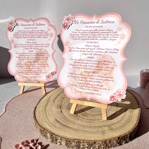 Può includere: Due menu con bordi smerlati intitolati La Comunione di Ludovica, decorati con illustrazioni floreali rosa e un'immagine centrale di una bambina. I cartoncini sono esposti su piccoli cavalletti in legno, uno su una fetta di legno.