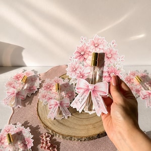 Può includere: Una piccola boccetta di profumo in vetro trasparente con erogatore dorato, fissata su un cartoncino decorativo con illustrazione di fiori rosa e un fiocco rosa. Il fiocco riporta la scritta "La mia Comunione" e il nome "Ludovica".