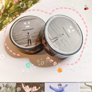 Könnte beinhalten: Zwei Rollen dekoratives Washi-Tape. Jede Rolle zeigt ein Schwarz-Weiß-Bild einer Person mit ausgestreckten Armen. Das Klebeband hat Text in einer Fremdsprache. Der Hintergrund ist weiß mit rosa gepunkteten Linien und floralen Akzenten.