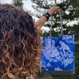 May include: A blue tote bag with a white toile pattern featuring figures and floral elements. The bag has gray denim accents at the bottom and gray straps. The bag is held up against a background of green foliage.