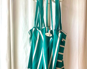 Puede incluir: Un bolso tote a rayas verdes y blancas con correas largas, colgado de un perchero blanco. El bolso tiene una pequeña etiqueta ovalada con texto. El bolso está hecho de tela y tiene un diseño sencillo e informal.