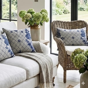 Könnte beinhalten: Ein gemütliches Wohnzimmer mit einem weißen Sofa, das mit Kissen mit blau-grauem Rautenmuster geschmückt ist. Eine gewebte Decke mit Fransen und ein Korbstuhl mit passendem Kissen sind zu sehen. Eine Vase mit grünen Hortensien fügt eine natürliche Note hinzu.