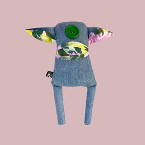 Può includere: Bambola in denim fatta a mano con accenti floreali sulle orecchie e sulla vita. La bambola ha un bottone verde e una piccola etichetta. Le gambe della bambola sono in denim.