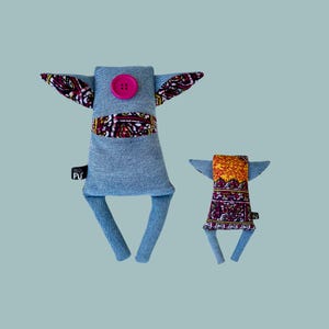 May include: Two handmade denim and fabric toys. The larger toy is blue denim with pink fabric ears and a pink button for a nose. The smaller toy is a multi-colored fabric with yellow, orange, and purple accents.