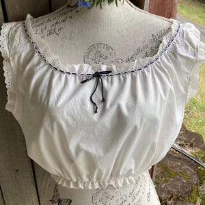 May include: A white, off-the-shoulder top with a ruffled neckline and hem. The top has eyelet lace detailing on the sleeves and a black bow. The top is displayed on a mannequin.