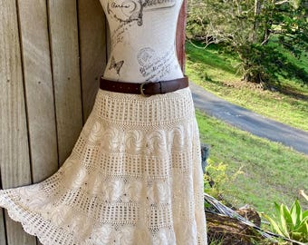 May include: A cream-colored, crochet skirt displayed on a mannequin. The skirt features a detailed, openwork design with a scalloped hem. A brown leather belt with a gold buckle cinches the waist. The mannequin is adorned with flowers.