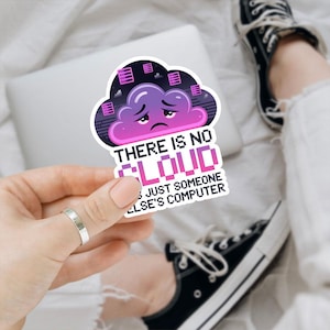 May include: A sticker featuring a purple cloud with a sad face and the text "THERE IS NO CLOUD JUST SOMEONE ELSE'S COMPUTER". The sticker has a pixelated design and is held by a hand. The background includes a laptop and a person's legs.