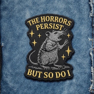 May include: Embroidered patch featuring a gray rat with an open mouth and outstretched arm. The black patch has gold text that reads "THE HORRORS PERSIST BUT SO DO I" with gold star accents. The patch is on a blue denim background.