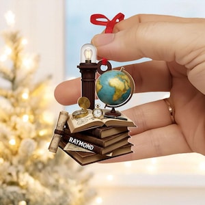 May include: A Christmas ornament featuring a miniature stack of books, a globe, a compass, and a small lightbulb. The ornament has a red ribbon for hanging and the name "Raymond" is printed on the books.