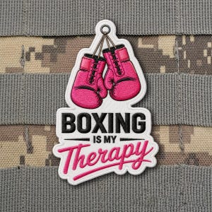 May include: A white patch with the text "BOXING IS MY Therapy" in black and pink lettering. The patch features a pair of pink boxing gloves hanging from a ring. The background is a camouflage pattern.