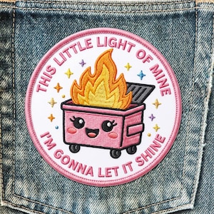 May include: A round, embroidered patch with the text "THIS LITTLE LIGHT OF MINE I'M GONNA LET IT SHINE" encircling a pink dumpster on wheels with flames. The patch is on a denim background, with star accents.