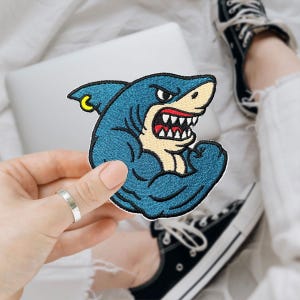 May include: Embroidered patch featuring a muscular shark with a fierce expression. The shark is blue with white teeth, a red mouth, and a yellow earring. The patch is held in front of a laptop and a person's legs.