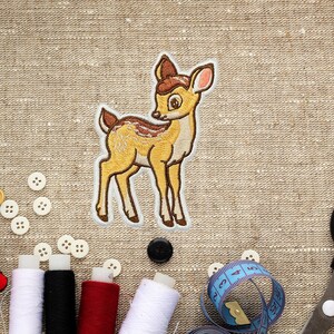 May include: Embroidered patch featuring a cartoon deer in shades of yellow, brown, and white. The deer is standing with its head turned to the side. Sewing supplies, including spools of thread, buttons, and a measuring tape, are visible around the patch.
