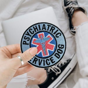 May include: A round patch with the words "PSYCHIATRIC SERVICE DOG" in blue lettering. The patch features a red star with a blue caduceus symbol in the center. The patch is being held by a person.