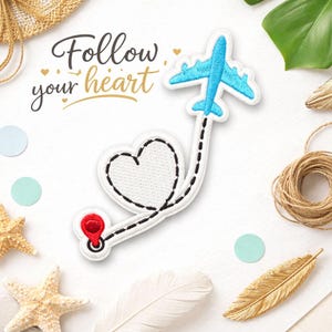 May include: A white patch with a black outline of a heart and a blue airplane, connected by a dotted line. The patch has a red pin at the end of the line. The text "Follow your heart" is written in black and gold.