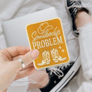 May include: Mustard yellow patch with the text "Somebody's PROBLEM" in white script. The patch features a cowboy hat and two cowboy boots. The patch is rectangular with a gold border.