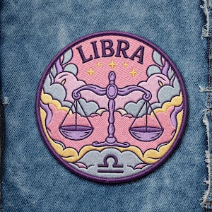 May include: Embroidered patch featuring the Libra zodiac symbol. The design includes a scale, stars, and the word "LIBRA" in a pink circle with a purple border. The patch is set against a blue denim background.