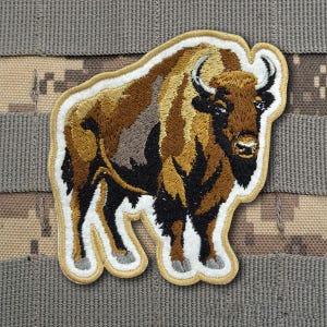 May include: An embroidered patch featuring a brown and tan American bison standing in profile. The patch has a thick, light tan border and white stitching outlining the animal's form, with detailed black and brown thread work depicting the fur.