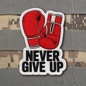 May include: A patch featuring two red boxing gloves with white trim. Below the gloves, the words "NEVER GIVE UP" are displayed in bold, black letters with a white border. The patch is set against a camouflage background.