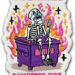 May include: Embroidered patch featuring a skeleton crying inside a purple dumpster engulfed in flames. The text "THE EMOTIONAL" and "DUMPSTER FIRE" are in pink. Black crosses and yellow stars surround the scene.