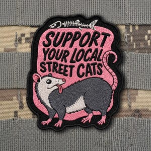 May include: Embroidered patch with a pink background and the text "SUPPORT YOUR LOCAL STREET CATS." A cartoon opossum with its tongue out is featured. A fish skeleton is at the top. The patch has a black border and is on a camouflage background.