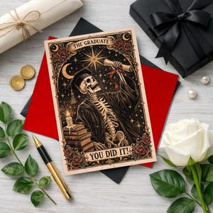 May include: A graduation card with a skeleton in a cap and gown holding a scroll, with the text "THE GRADUATE" and "YOU DID IT!". The card is surrounded by roses, a candle, and a starry night sky. The card is on a red and black background.