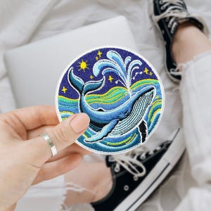 May include: Embroidered patch featuring a blue whale breaching the ocean's surface. The round patch has a dark blue background with yellow stars and a sun. The whale is detailed with white lines, and the waves are blue and green.