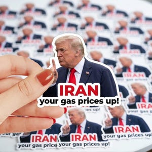 May include: A sticker featuring a portrait of a man in a suit and red tie, with the text "IRAN your gas prices up!" The background shows multiple stickers with the same image. The sticker is held by a hand with a red nail polish.