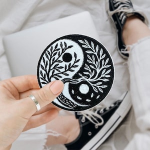 May include: A round black and white Yin Yang patch with a tree design. The patch is held in a hand with a silver ring. A laptop and black sneakers are in the background.