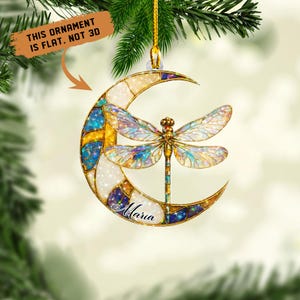 May include: A decorative ornament featuring a dragonfly with iridescent wings, set against a crescent moon. The moon is segmented with white, gold, blue, and purple sections. The name "Maria" is written on the moon. The text "THIS ORNAMENT IS FLAT. NOT 3D" is also present.