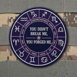May include: A circular patch with a navy blue background and a white and pink border. The patch features zodiac symbols around the edge and the text "YOU DIDN'T BREAK ME, YOU FORGED ME." in white. The patch is on a camouflage background.