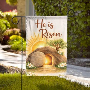 May include: A garden flag with the text "He is Risen" in brown script. The flag features a watercolor illustration of an open tomb with a rising sun, a tree, and white flowers. The flag is hanging on a black metal stand.