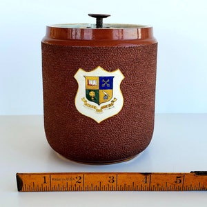 Puede incluir: Un tarro de cerámica marrón con tapa y un escudo que presenta un libro, llaves cruzadas, un árbol, un pájaro y el texto "Disce, Doce, Doctus" y "Cheltenham College Training".