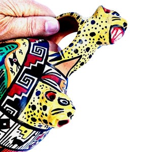 Puede incluir: Un recipiente de cerámica pintado a mano con un pico y asa con forma de cabeza de jaguar. El recipiente presenta una cabeza de jaguar amarilla con manchas negras y detalles en rojo. El cuerpo del recipiente está decorado con patrones geométricos en rojo, negro, azul y amarillo.