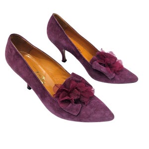 May include: A pair of deep purple suede pumps featuring a pointed toe and a kitten heel. Each shoe is adorned with a layered, ruffled fabric flower in a matching dark plum shade on the vamp. The interior lining is a tan leather with gold branding.