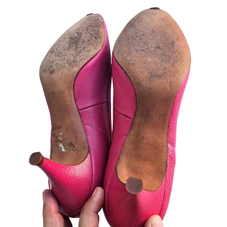 May include: Pair of bright pink leather heels with pointed toes and small heels. The soles are worn, showing signs of use. The shoes are held up by a hand against a white background.