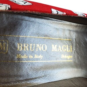 May include: Close-up of the inside of a shoe with the brand name "BRUNO MAGLI" in gold lettering. The shoe's interior is a dark gray color, and the red fabric trim has a white and black pattern.