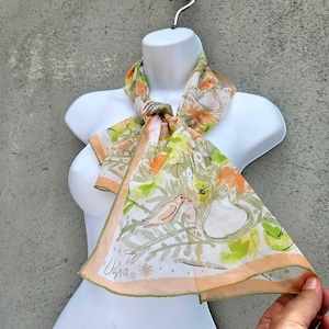 May include: A lightweight scarf featuring a white background with a watercolor-style print of orange birds, green foliage, and abstract shapes. The scarf has a light orange border and includes the signature text "Usna" printed in the bottom corner.