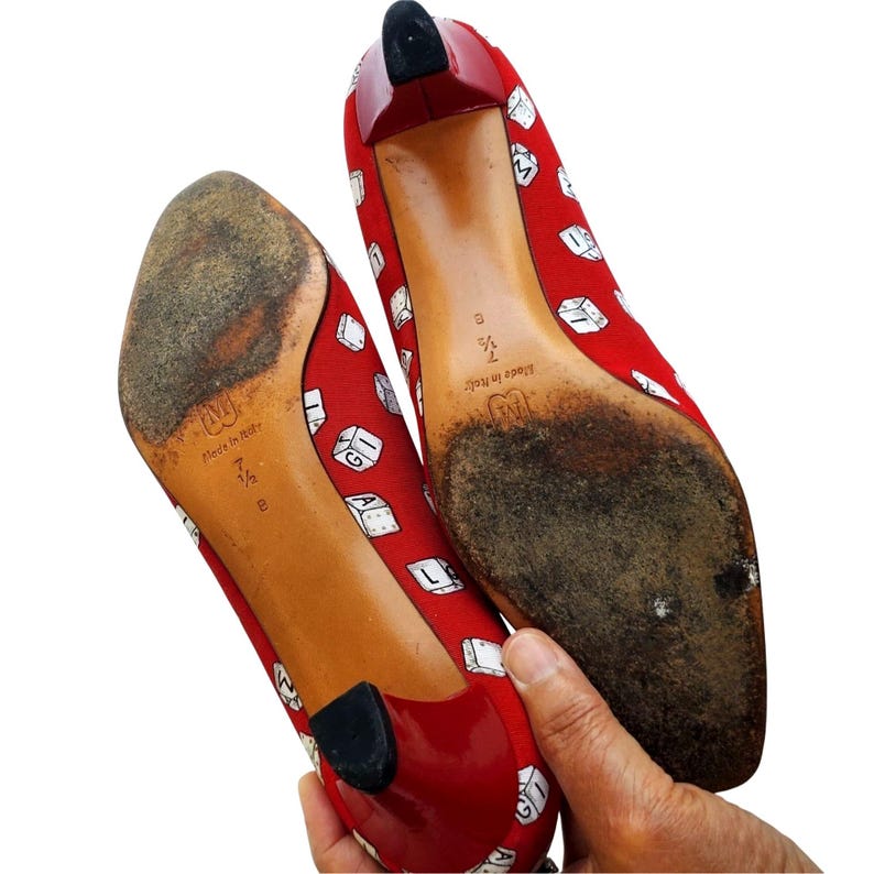May include: A pair of red high heels with a dice pattern. The heels have a red heel and a black tip. The soles are worn and show the size 7 1/2. The shoes are held by a hand.