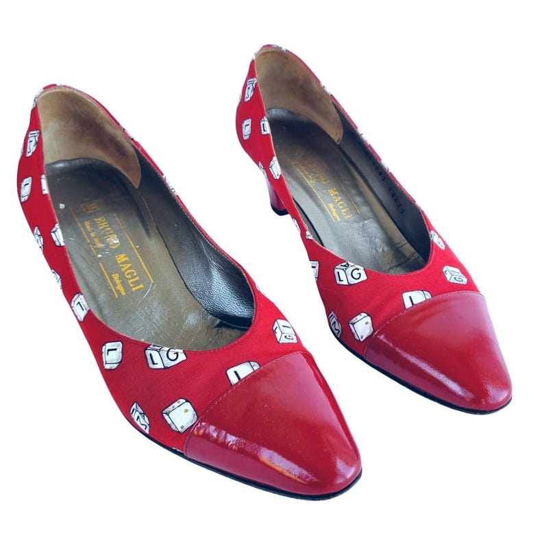 May include: A pair of red heels with a glossy finish on the toe and a fabric upper patterned with white dice. The heels have a low heel and the brand name "BRUNO MAGLI" is visible inside the shoe.